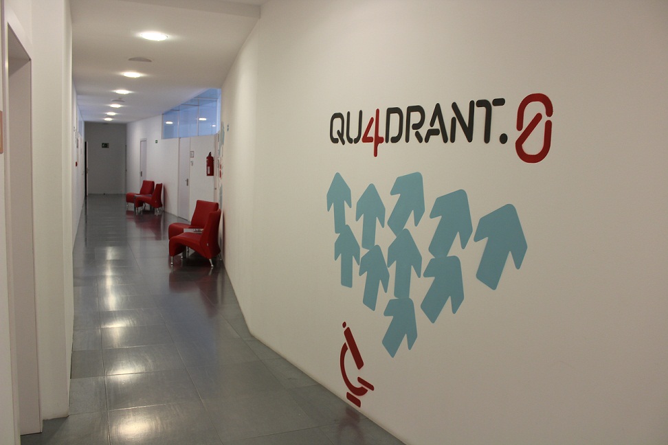Imatge passadís Qu4drant.0 on surten cadires, portes de sales i una imatge a la paret blanca amb fletxes blaves i el nom "Qu4drant.0"